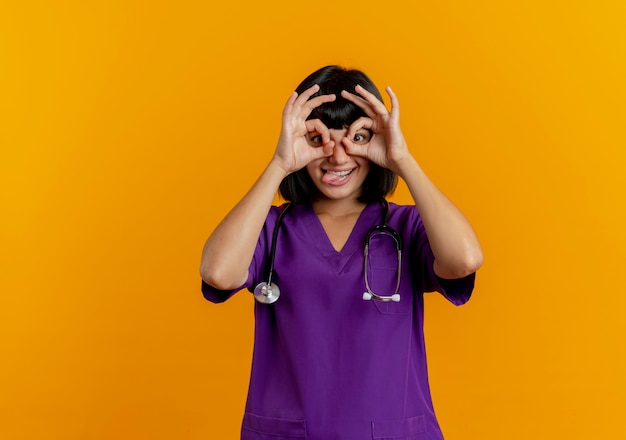 Médica jovem morena engraçada de uniforme com estetoscópio enfiando a língua para fora