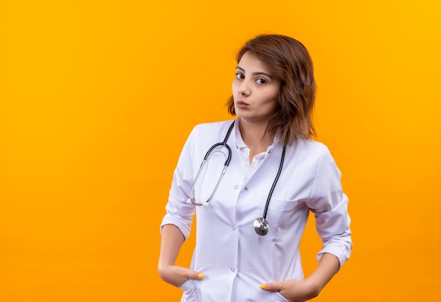 Médica jovem vestindo jaleco branco com estetoscópio em pé com as mãos nos bolsos e rosto sério na parede laranja