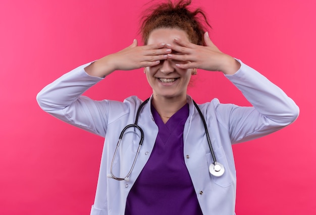 Médica jovem vestindo jaleco branco com estetoscópio fechando os olhos com os dedos sorrindo em pé sobre a parede rosa