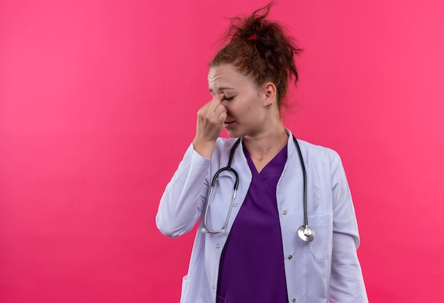 Médica jovem vestindo jaleco branco com estetoscópio, parecendo cansada e sobrecarregada, tocando o nariz entre os olhos fechados em pé sobre uma parede rosa