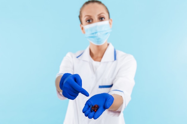 Médica loira linda usando uniforme e máscara em pé isolado sobre a parede azul, mostrando comprimidos na palma da mão