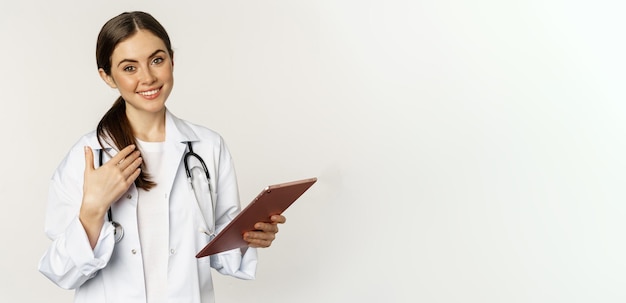 Foto grátis médica morena sorridente segurando tablet digital apontando para si mesma com cara amigável em pé