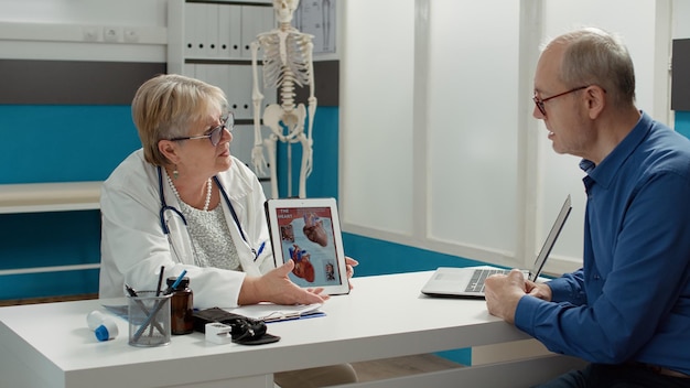 Médica mostrando diagnóstico de cardiologia no tablet digital, segurando o gadget com ilustração do sistema cardiovascular para aposentado. condição cardíaca e doença na visita de check-up.