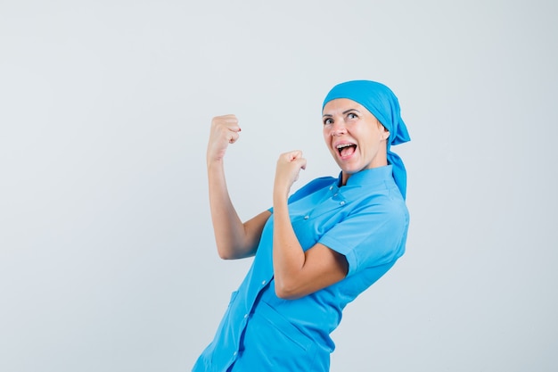 Médica mostrando gesto de vencedor em uniforme azul e olhando feliz, vista frontal.