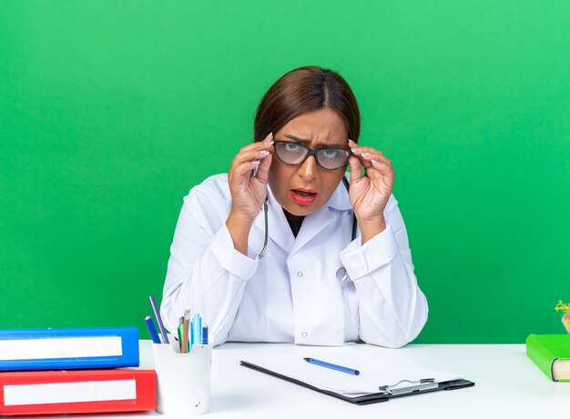 Foto grátis médica mulher de jaleco branco com estetoscópio e óculos confusa e muito ansiosa sentada à mesa sobre a parede verde