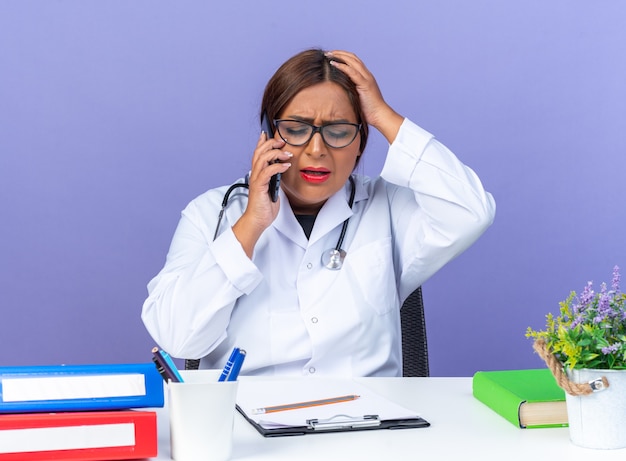 Médica mulher de jaleco branco com estetoscópio e óculos, parecendo confusa enquanto fala ao telefone celular, sentada à mesa sobre a parede azul