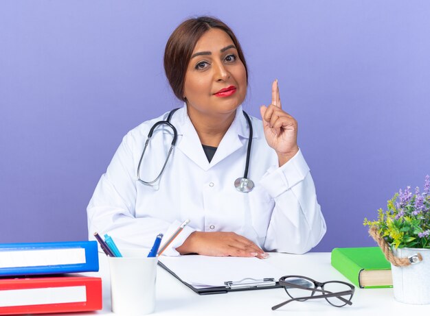 Médica mulher de jaleco branco com estetoscópio olhando com expressão confiante, mostrando o dedo indicador como um aviso, sentada à mesa sobre fundo azul