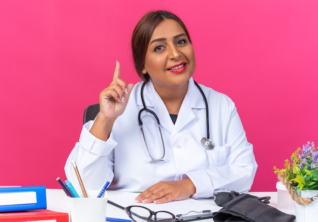 Médica mulher de jaleco branco com estetoscópio olhando sorrindo alegremente, mostrando o dedo indicador tendo uma nova ideia sentada à mesa com pastas de escritório sobre fundo rosa