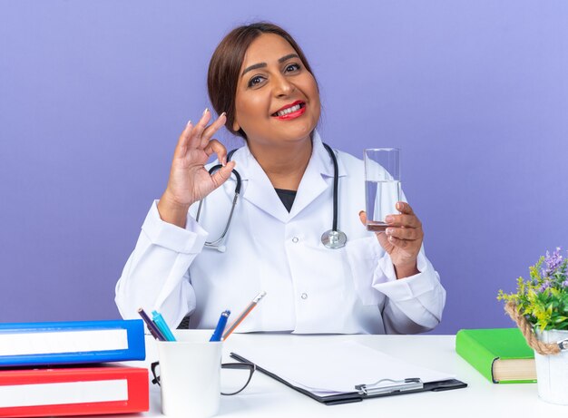 Médica mulher de jaleco branco com estetoscópio segurando um copo de água, olhando para a frente feliz e sorrindo positivamente fazendo sinal de ok sentado à mesa sobre a parede azul