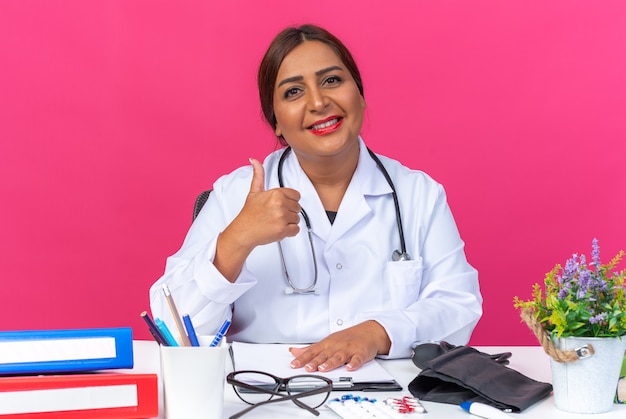 Médica mulher de meia-idade vestindo jaleco branco com estetoscópio sorrindo confiante, mostrando os polegares para cima, sentada à mesa com pastas de escritório rosa