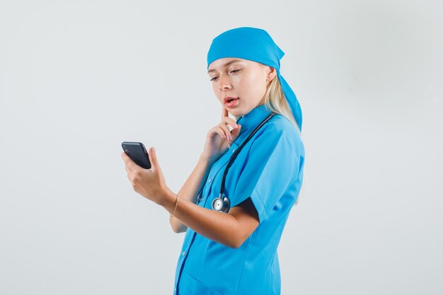 Médica pensando enquanto olha para um smartphone de uniforme azul