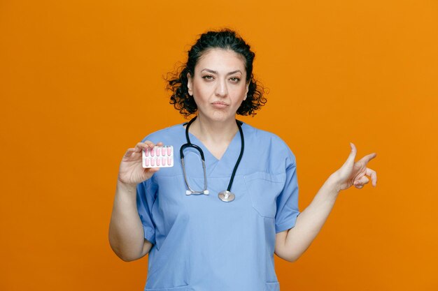 Médica séria de meia-idade vestindo uniforme e estetoscópio em volta do pescoço, mostrando o pacote de cápsulas olhando para a câmera apontando para o lado isolado em fundo laranja