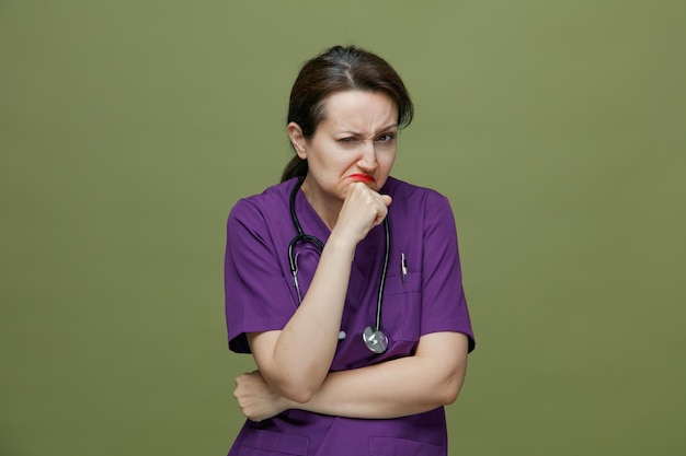 Médica suspeita de meia-idade vestindo uniforme e estetoscópio no pescoço, mantendo a mão sob o cotovelo outra mão no queixo olhando para câmera isolada em fundo verde oliva