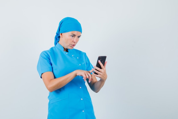 Médica usando telefone celular com uniforme azul e parecendo surpresa. vista frontal.