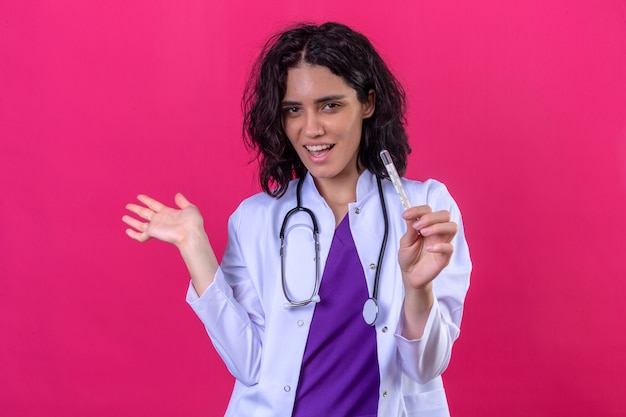 médica vestindo jaleco branco com estetoscópio mostrando o termômetro apontando com a mão para o lado com um sorriso no rosto de pé na rosa isolada
