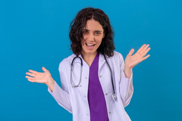 médica vestindo jaleco branco com estetoscópio olhando surpresa levantando as palmas das mãos sorrindo no isolado azul