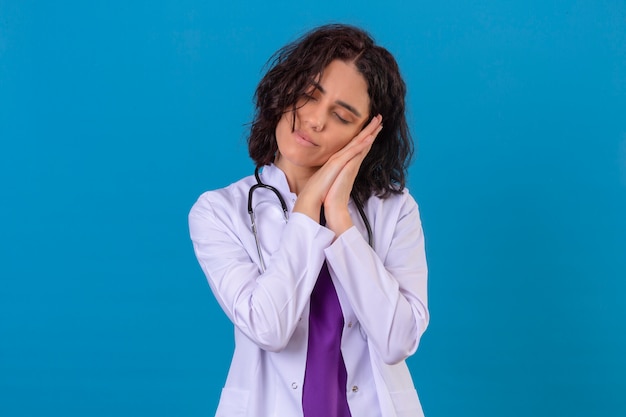 médica vestindo jaleco branco com estetoscópio, parecendo um trabalho, posando com as mãos juntas enquanto sorri com os olhos fechados, quer dormir no isolado azul