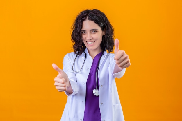 médica vestindo jaleco branco com estetoscópio sorrindo alegre mostrando os polegares para cima em laranja isolada