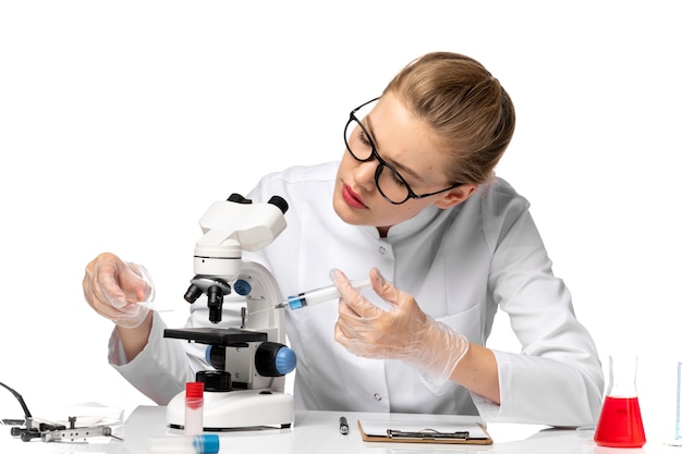 Médica vista frontal em traje médico branco com luvas segurando a injeção e usando microscópio no espaço em branco