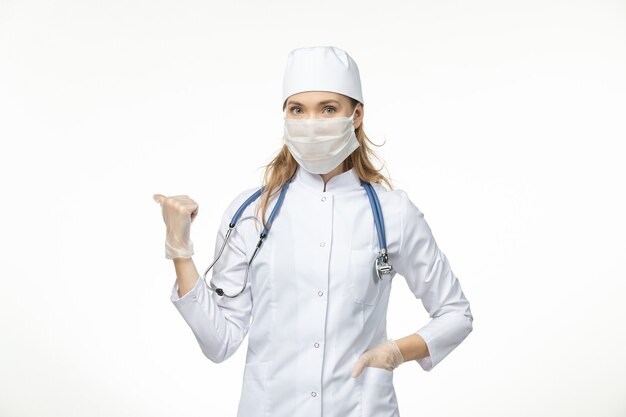 Médica vista frontal em traje médico com máscara estéril devido a coronavírus na doença da parede branca, doença pandêmica covid-