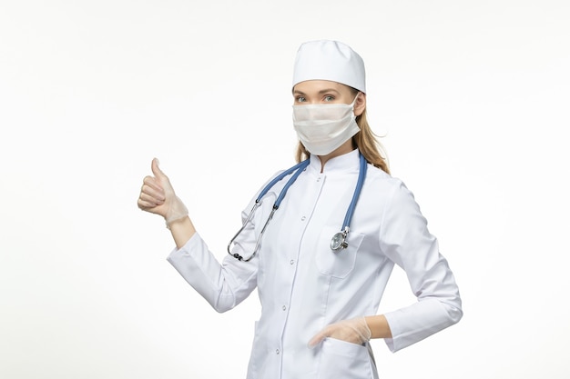 Médica vista frontal em traje médico usando máscara devido a coronavírus na doença de mesa branca clara vírus covidpandêmico