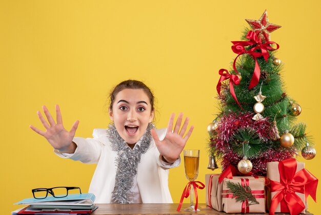 Médica vista frontal sentada ao redor de uma árvore e presentes de Natal em fundo amarelo