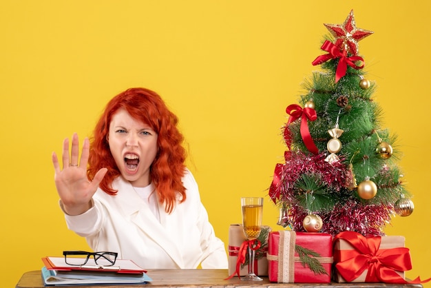 Médica vista frontal sentada atrás da mesa com os presentes de Natal em fundo amarelo