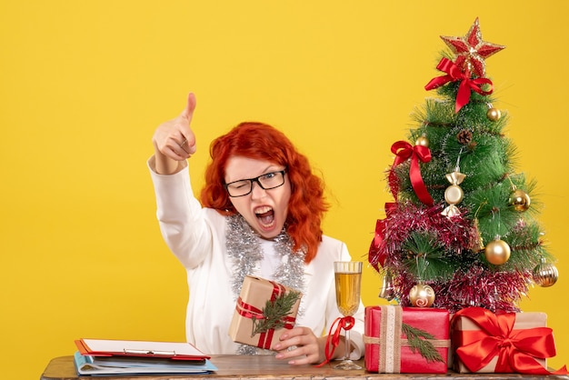 Médica vista frontal sentada atrás da mesa com os presentes de natal em fundo amarelo