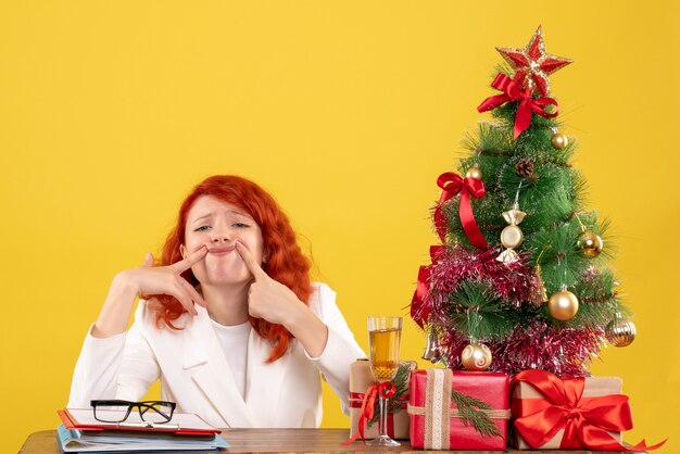 Médica vista frontal sentada atrás da mesa com os presentes de Natal em fundo amarelo