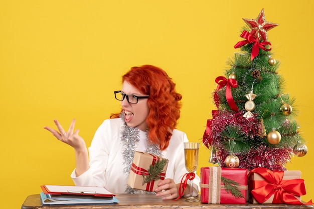 Médica vista frontal sentada atrás da mesa com presentes de natal em um fundo amarelo com árvore de natal e caixas de presente