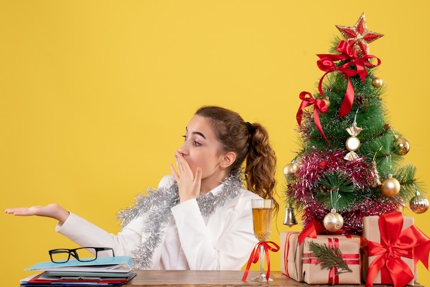 Médica vista frontal sentada atrás da mesa em fundo amarelo com árvore de Natal e caixas de presente