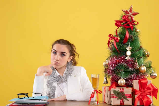 Médica vista frontal sentada atrás da mesa pensando no fundo amarelo com árvore de Natal e caixas de presente