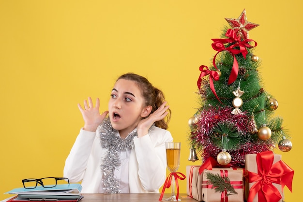 Médica vista frontal sentada atrás de sua mesa com cara assustada em fundo amarelo com árvore de Natal e caixas de presente