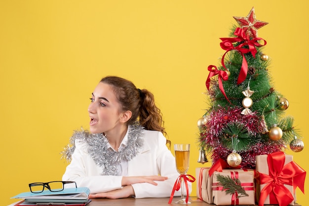 Médica vista frontal sentada atrás de sua mesa em fundo amarelo com árvore de Natal e caixas de presente