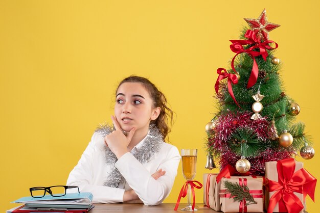 Médica vista frontal sentada atrás de sua mesa em fundo amarelo com árvore de natal e caixas de presente