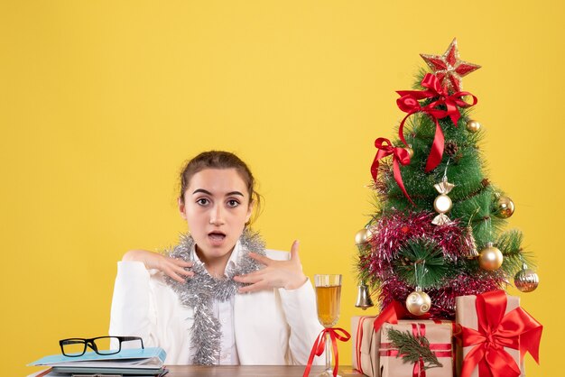 Médica vista frontal sentada atrás de uma mesa com presentes de Natal e árvore no fundo amarelo