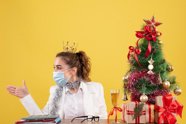Médica vista frontal sentada com máscara estéril, apertando as mãos em fundo amarelo com árvore de natal e caixas de presente