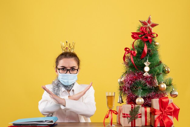 Médica vista frontal sentada com máscara estéril em fundo amarelo com árvore de natal e caixas de presente