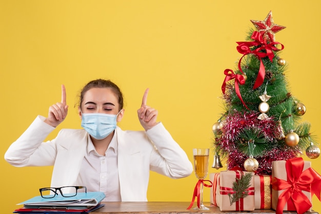 Médica vista frontal sentada com máscara protetora em fundo amarelo com árvore de natal e caixas de presente