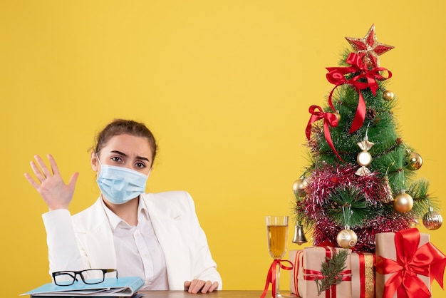 Médica vista frontal sentada com máscara protetora em fundo amarelo com árvore de natal e caixas de presente