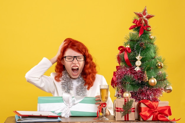 Médica vista frontal sentada com presentes de natal e árvore em fundo amarelo