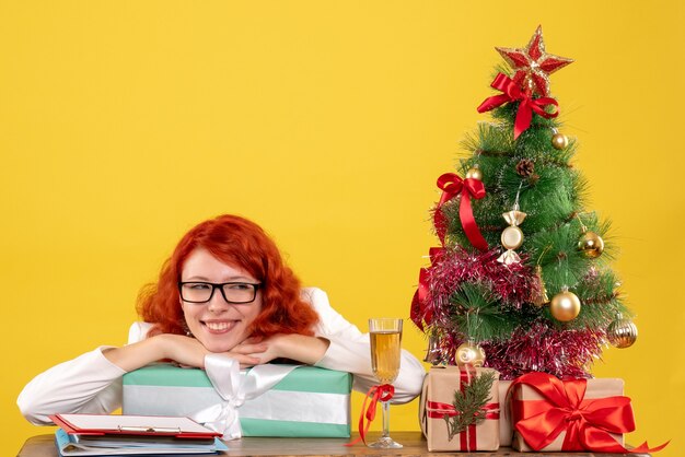 Médica vista frontal sentada com presentes de Natal e árvore em fundo amarelo