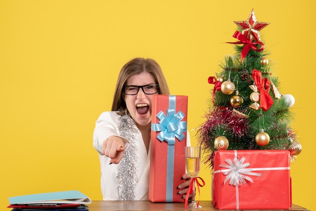 Médica vista frontal sentada com presentes de natal e árvore no fundo amarelo