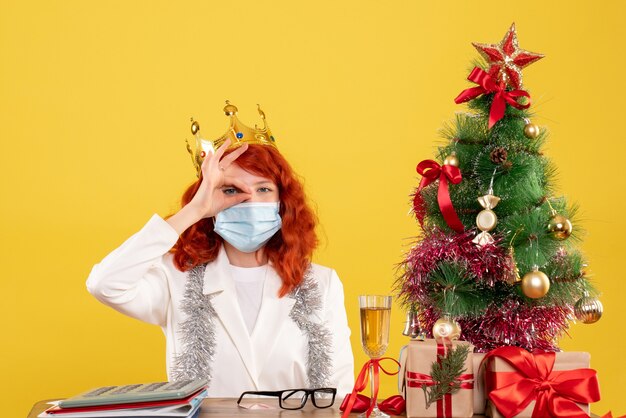 Médica vista frontal sentada com presentes de natal e usando coroa sobre fundo amarelo