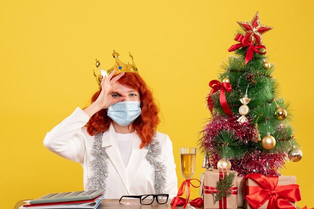 Médica vista frontal sentada com presentes de Natal e usando coroa sobre fundo amarelo