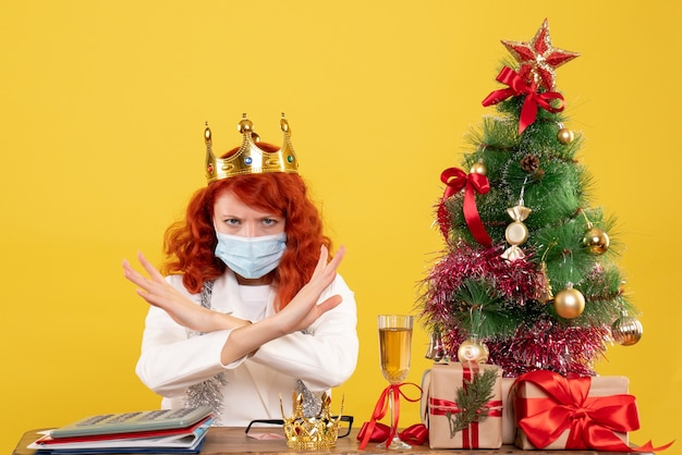 Médica vista frontal sentada com presentes de natal e usando coroa sobre fundo amarelo