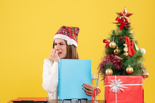 Médica vista frontal sentada com presentes de Natal segurando arquivos em fundo amarelo