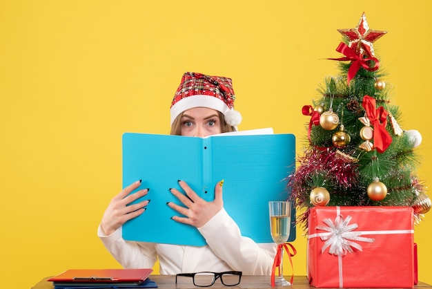 Médica vista frontal sentada com presentes de natal segurando arquivos em fundo amarelo