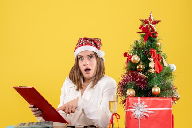 Médica vista frontal sentada com presentes de Natal segurando uma nota de arquivo sobre fundo amarelo