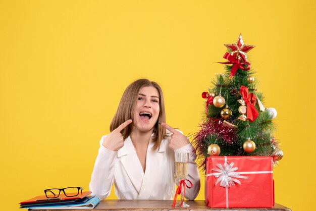 Médica vista frontal sentada em frente à mesa com presentes de Natal e árvore no fundo amarelo